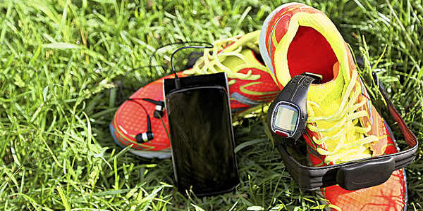 Black sports watch, smartphone and headphones placed on a pair of yellow and red sneakers on the grass.