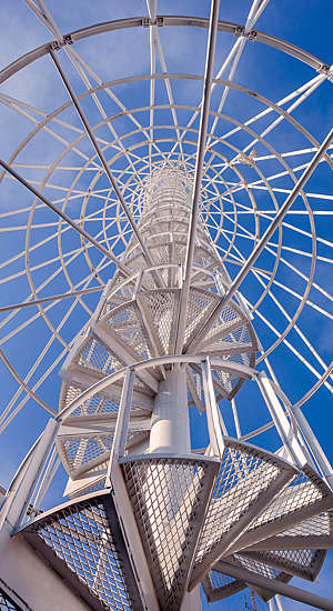 Low-angle shot of a white-metal telephone communication tower with spiral staircase, photographed against the blue sky.