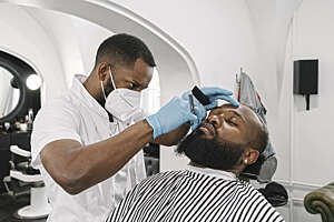 Lithuania, Vilnius, Barber wearing face mask and reusable  gloves doing beard of customer in a barber shop